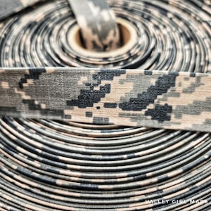 May include: Close-up of a roll of camouflage ribbon. The ribbon features a digital camouflage pattern in shades of tan, gray, and black. The ribbon is tightly wound, revealing the edges of each layer. The text "VALLEY GIRL MADE" is visible at the bottom.