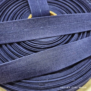 May include: A roll of navy blue bias tape, with a visible texture and a folded edge. The tape is neatly coiled, with two lengths of tape laid across the top. The text "VALLEY GIRL MADE" is visible in the bottom right corner.