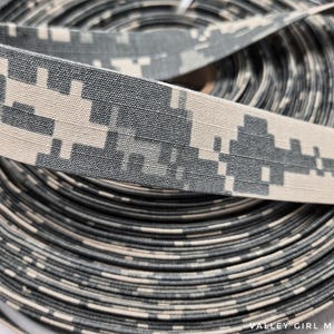 May include: A roll of digital camouflage ribbon. The ribbon features a pixelated pattern in shades of gray, tan, and black. The ribbon is tightly wound, with the top layer showing the detailed pattern. The text "VALLEY GIRL MADE" is visible at the bottom.