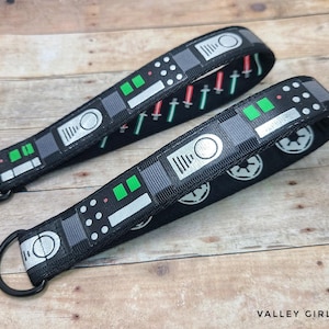 May include: Two black key fobs with a repeating pattern of gray and white geometric shapes, green and red accents, and a black metal ring. The design includes images of lightsabers and the Imperial symbol.