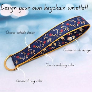 May include: A navy blue keychain wristlet with a gold-colored D-ring. The wristlet features a design with red roses and gold accents. Text on the image reads "Design your own keychain wristlet!" and prompts to choose design and color.