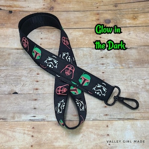 May include: Black lanyard with a repeating pattern of Star Wars characters, including Darth Vader, Boba Fett, and Stormtroopers. The lanyard is glow-in-the-dark and has a black metal clip. The text "Glow in the Dark" is written in green on a white background.