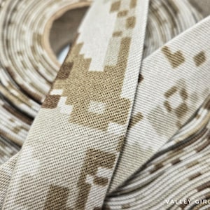 May include: Close-up of a roll of fabric with a digital camouflage pattern in shades of tan, brown, and beige. The fabric is tightly rolled, with the pattern visible on the edges and the surface. The text "VALLEY GIRL MADE" is visible in the bottom right corner.