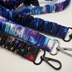 May include: Three fabric lanyards with metal swivel hooks. The lanyards feature a variety of space-themed patterns, including a black one with colorful stars, and two with blue and purple galaxy designs.