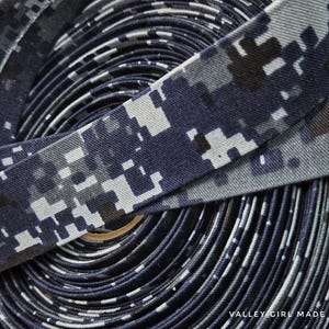 May include: Close-up of a roll of navy blue and gray digital camouflage fabric. The fabric is tightly wound, revealing a pattern of small, square shapes in varying shades of blue, gray, and white. The fabric is likely used for sewing or crafting projects.