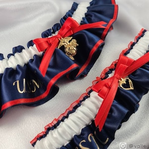May include: Two blue and white garters with red ribbon bows. One garter has a gold charm with the USMC logo and the other has a gold heart charm. The garter with the USMC logo has the letters "USMC" embroidered in gold on the blue satin.