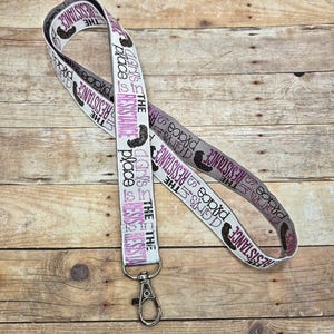 May include: A white, gray, and pink lanyard with the text "THE RESISTANCE" and "A Girl's Place is the Resistance" printed on it. The lanyard has a silver-colored metal clasp.