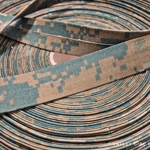 May include: A roll of fabric trim with a digital camouflage pattern in shades of tan and green. The trim is tightly wound, revealing the pattern's repeating design. The fabric appears to be a sturdy material, suitable for various crafting projects.