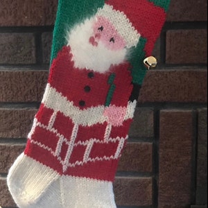 May include: A red, white, and green knitted Christmas stocking with a Santa Claus design and the name "Dylan" embroidered on the top. The stocking has a bell attached to the top.