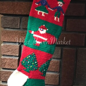 May include: A red and green knitted Christmas stocking with the name "Piper" embroidered on the top. The stocking features a Santa Claus, two dancing figures, and a Christmas tree with colorful beads.