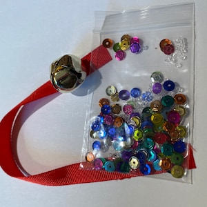 May include: A red ribbon with a silver bell attached to one end. A clear plastic bag filled with colorful sequins and clear beads is attached to the other end of the ribbon.