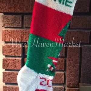 May include: A green, red, and white knitted Christmas stocking with the name "Winnie" embroidered on the front. The stocking has a small bell at the bottom and a Christmas tree design with ornaments.