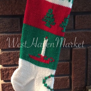 May include: A knitted Christmas stocking with the name "MELIN" in green letters on a white background. The stocking features red, green, and white sections with Christmas tree and candle designs. A gold bell hangs from the top.