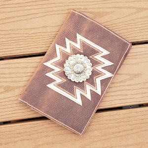 May include: A brown leather wallet with a white geometric pattern and a silver concho. The wallet has a distressed look.