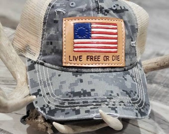 May include: A camouflage trucker hat with a brown leather patch featuring a red, white, and blue American flag and the text "Live Free or Die".