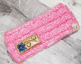 May include: Pink knit headband with a brown leather patch featuring a blue flower and green leaves. The headband has a fluffy lining and the leather patch has the text "C.C. Kids".