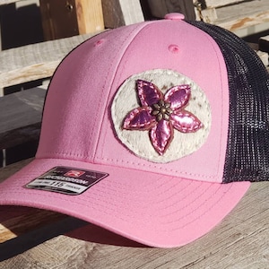 Peut inclure: Casquette de camionneur rose et noire avec un patch de fleur blanche et rose. Le patch a un bouton marron au centre.