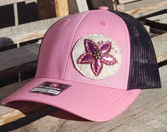 May include: Pink and black trucker hat with a white and pink flower patch. The patch has a brown button in the center.