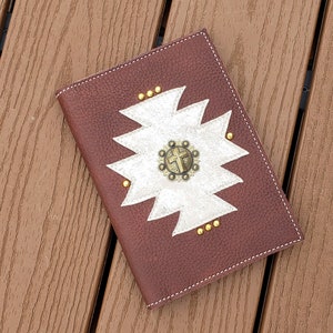 May include: A brown leather journal with a white and silver geometric design and a gold cross accent. The journal has a stitched edge and is closed.