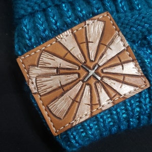 May include: A teal blue knitted beanie with a brown leather patch featuring a white windmill design.