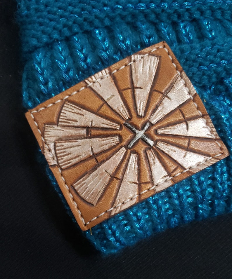 May include: A teal blue knitted beanie with a brown leather patch featuring a white windmill design.