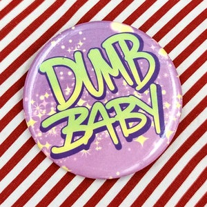 May include: A purple button with the words "DUMB BABY" written in green and yellow letters with a glittery background.