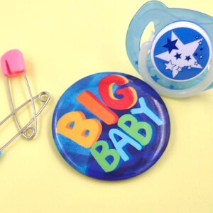 May include: A blue and white pacifier with a star design and a blue button with the words "BIG BABY" in colorful letters.