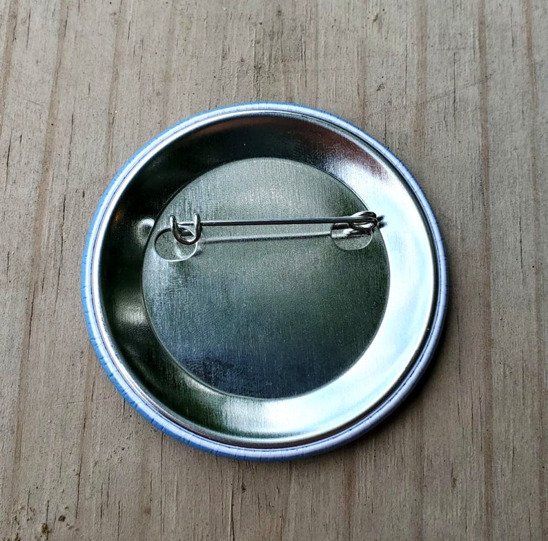 May include: The back of a round button with a blue border. The button has a metal backing with a safety pin attached.