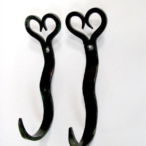 May include: Two black metal wall hooks with a heart-shaped top design. The hooks have a curved, elongated body and a curved hook at the bottom. The hooks are made of a dark, possibly wrought iron material.