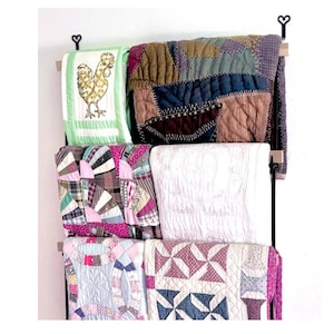 May include: A black metal quilt rack with four quilts hanging on it. The quilts are in various patterns and colors, including pink, green, brown, and white. The rack has two wooden dowels and heart-shaped hooks.