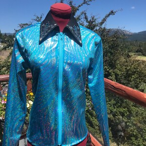 May include: A long-sleeved, turquoise jacket with a shimmering, iridescent pattern. The jacket features a black collar with decorative accents and a full-length zipper. The garment is displayed on a mannequin against a backdrop of a blue sky and greenery.