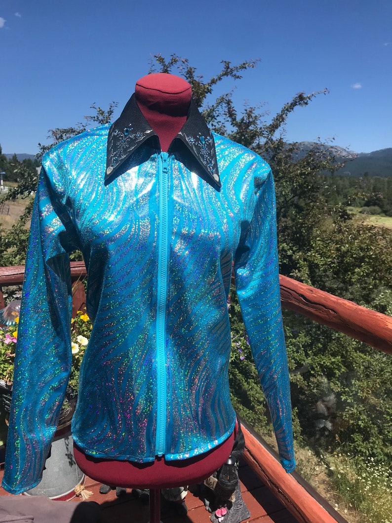 May include: A long-sleeved, turquoise jacket with a shimmering, iridescent pattern. The jacket features a black collar with decorative accents and a full-length zipper. The garment is displayed on a mannequin against a backdrop of a blue sky and greenery.