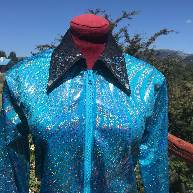 May include: A turquoise, shimmering jacket with a zebra stripe pattern. The jacket has a black collar with silver and blue embellishments and a full-length zipper. The jacket is displayed on a mannequin against a backdrop of trees and a blue sky.