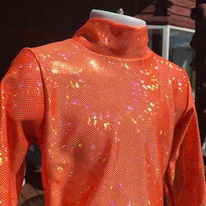 May include: Orange sequin turtleneck top. The long-sleeved top features a high, close-fitting collar and is made of a shiny, perforated fabric. The sequins reflect light, creating a sparkling effect. The top is displayed on a mannequin.