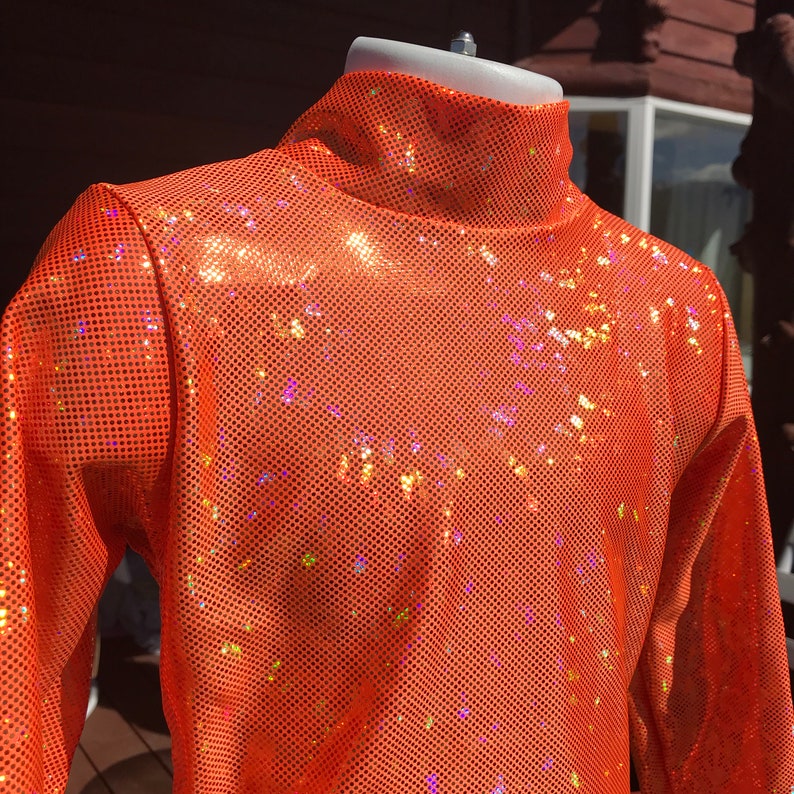 May include: Orange sequin turtleneck top. The long-sleeved top features a high, close-fitting collar and is made of a shiny, perforated fabric. The sequins reflect light, creating a sparkling effect. The top is displayed on a mannequin.