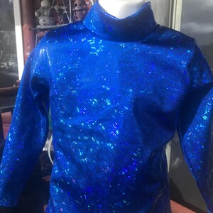 May include: A bright blue, long-sleeved top with a high, roll neck. The fabric has a shimmering, textured appearance, with a pattern of small, reflective squares. The top is displayed on a white mannequin, showcasing its form and design. The overall style suggests a performance or costume piece.