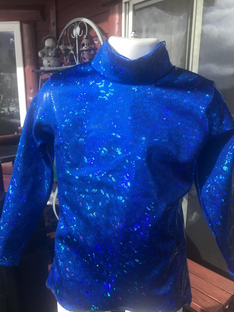 May include: A bright blue, long-sleeved top with a high, roll neck. The fabric has a shimmering, textured appearance, with a pattern of small, reflective squares. The top is displayed on a white mannequin, showcasing its form and design. The overall style suggests a performance or costume piece.
