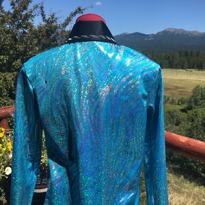 May include: A turquoise, long-sleeved jacket with a shimmering, wave-like pattern. The jacket has a black collar with white trim and a red and black hat. The background features a scenic view of mountains and trees.