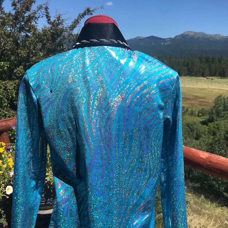 May include: A turquoise, long-sleeved jacket with a shimmering, wave-like pattern. The jacket has a black collar with white trim and a red and black hat. The background features a scenic view of mountains and trees.