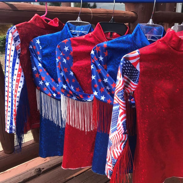 May include: Several red, white, and blue patriotic outfits hang on a blue bar. The outfits have long sleeves and are decorated with stars, stripes, and fringe. The outfits are likely for a performance or celebration.