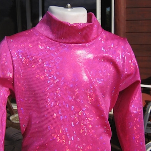 May include: A vibrant pink, long-sleeved turtleneck top. The fabric has a textured, shimmering appearance with a grid-like pattern. The top is displayed on a mannequin.