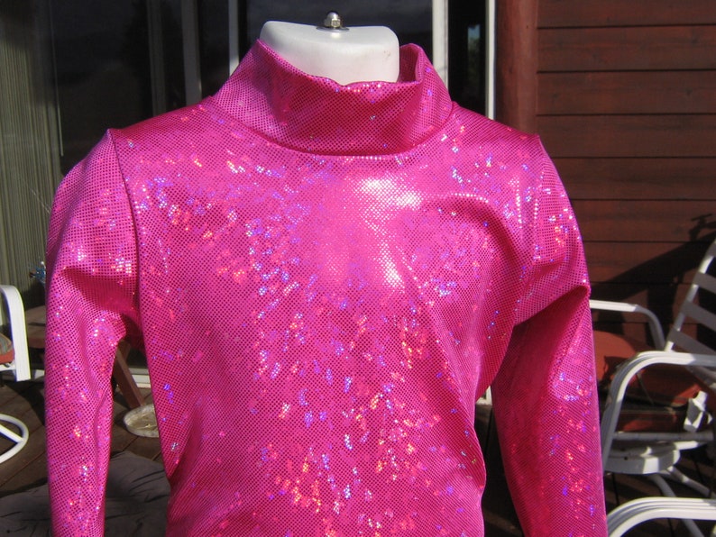May include: A vibrant pink, long-sleeved turtleneck top. The fabric has a textured, shimmering appearance with a grid-like pattern. The top is displayed on a mannequin.