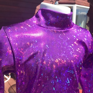 May include: A bright purple, long-sleeved turtleneck top. The fabric has a textured, sequined appearance, catching light with iridescent flashes of colour. The top is displayed on a white mannequin head.