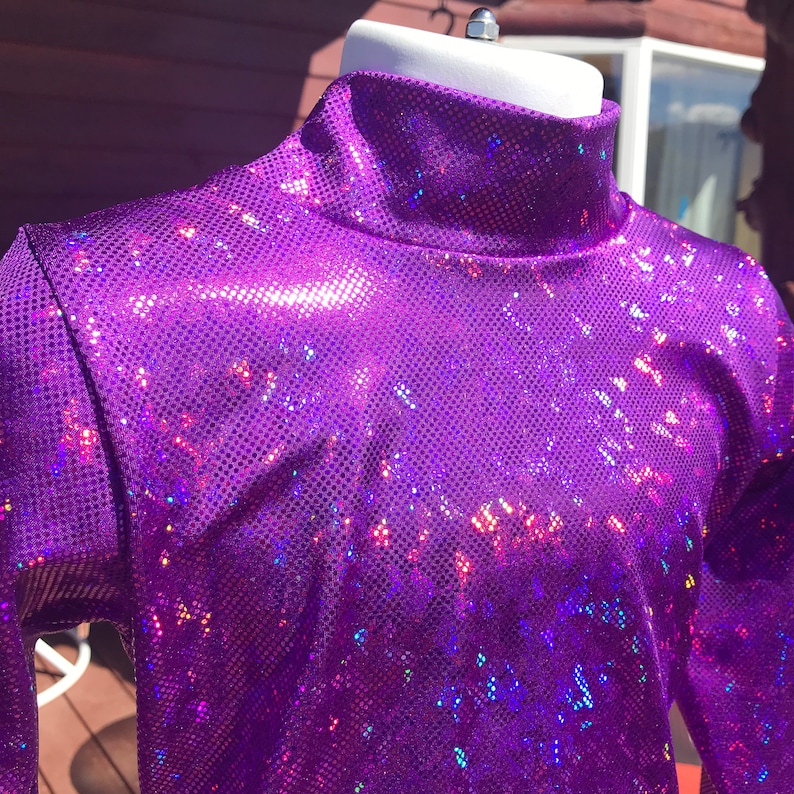 May include: A bright purple, long-sleeved turtleneck top. The fabric has a textured, sequined appearance, catching light with iridescent flashes of colour. The top is displayed on a white mannequin head.