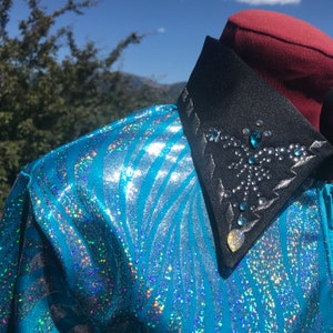 May include: A turquoise and silver equestrian show jacket with a black collar. The jacket has a shimmering, striped pattern. The collar is embellished with rhinestones and a decorative design.