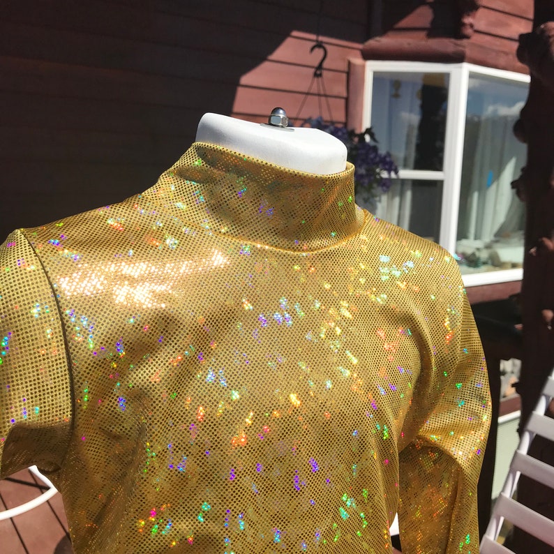 May include: A gold, long-sleeved turtleneck top with a shimmering, sequined finish. The fabric has a perforated texture and reflects multiple colours. The top is displayed on a white mannequin head.