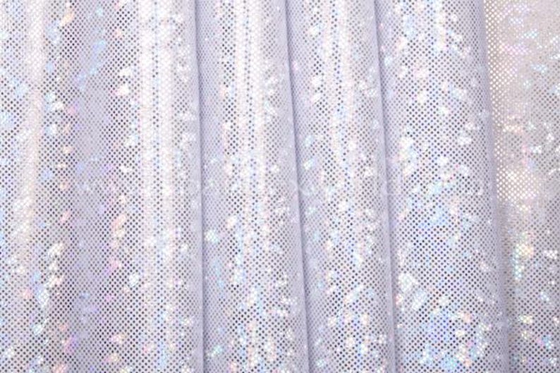 May include: Close-up of white fabric with a grid pattern and iridescent glitter accents. The fabric is draped in soft folds, creating a textured surface. The overall effect is a shimmering, light-reflecting material.
