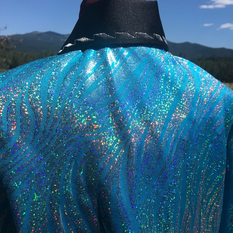 May include: A close-up of a turquoise jacket with a shimmering, iridescent pattern. The jacket has a black collar with white stitching. The fabric has a wavy, zebra-like design with sparkling accents.