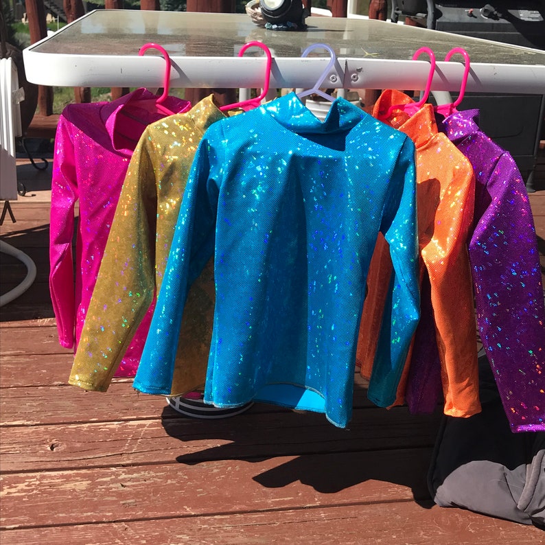 May include: Several long-sleeved tops in a range of colours: pink, gold, blue, orange, and purple. Each top features a high neckline and a shimmering, textured fabric. The tops are displayed on pink and purple hangers.