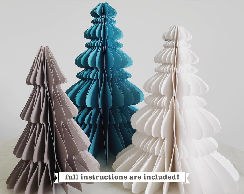 Puede incluir: Tres &aacute;rboles de Navidad de papel en diferentes tama&ntilde;os y colores: un &aacute;rbol gris, un &aacute;rbol azul verdoso y un &aacute;rbol blanco. El texto "&iexcl;Instrucciones completas incluidas!" est&aacute; en la parte inferior de la imagen.